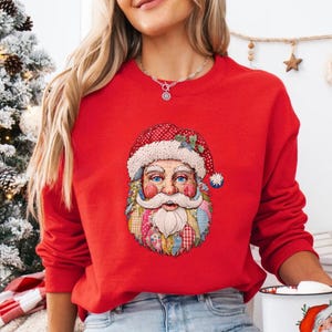 May include: A red sweatshirt featuring a colorful Santa Claus graphic. The Santa Claus design includes a red hat with white polka dots, a white beard, and a patchwork design on the face. The sweatshirt is a solid red color.