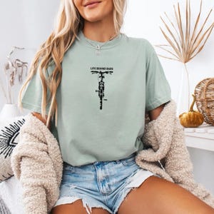 May include: A light green t-shirt with the text "LIFE BEHIND BARS" above a black bicycle graphic. The shirt is a relaxed fit and has short sleeves. The model is wearing denim shorts and a cream-colored cardigan.