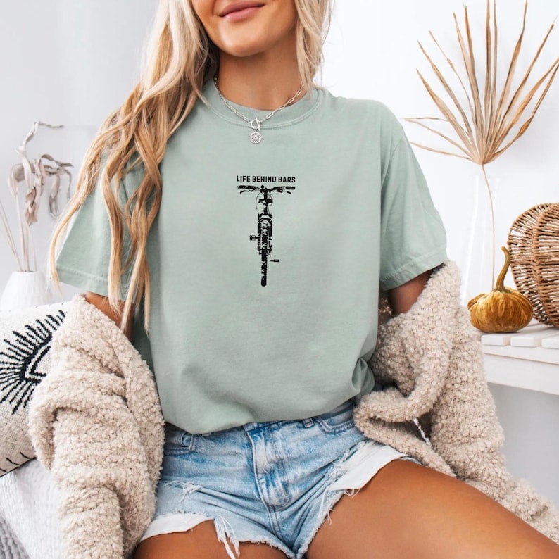 May include: A light green t-shirt with the text "LIFE BEHIND BARS" above a black bicycle graphic. The shirt is a relaxed fit and has short sleeves. The model is wearing denim shorts and a cream-colored cardigan.