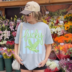 May include: A light blue t-shirt with the word "lucky" in green script and a green duck graphic. The shirt has blue vertical stripes behind the duck. The person is wearing a tan baseball cap and sunglasses.