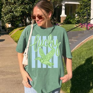 May include: Green t-shirt with the word "Lucky" in cursive script and a green duck graphic. The design features light blue vertical stripes. The shirt is a relaxed fit.