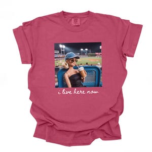 Pu&ograve; includere: T-shirt bordeaux con una grafica di una bambola con occhiali da sole e berretto da baseball, che tiene in mano una bevanda, con uno stadio di baseball sullo sfondo. Il testo "i live here now" &egrave; sotto la grafica.