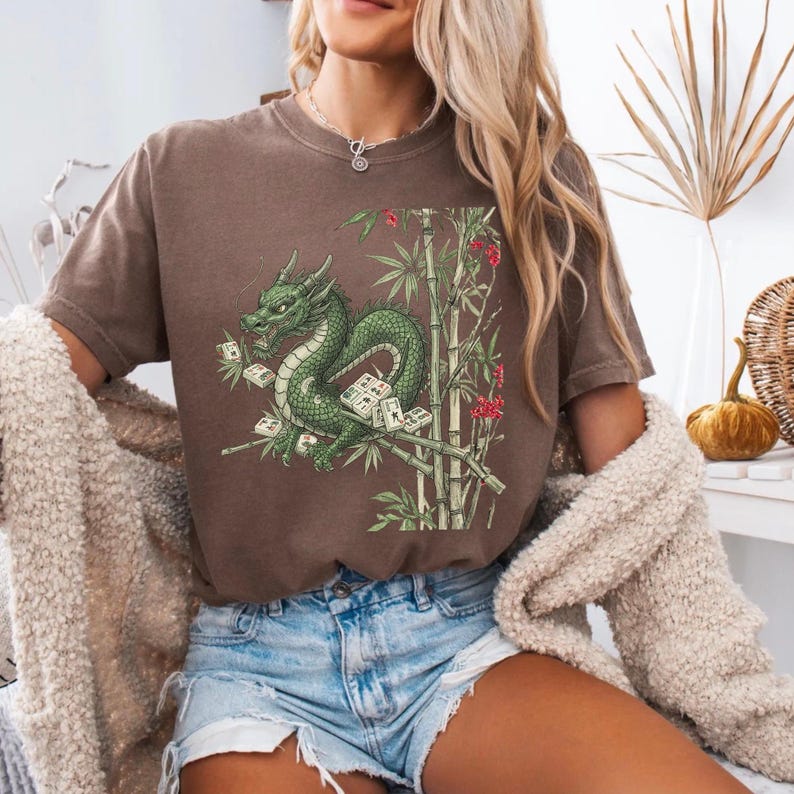 May include: Brown t-shirt featuring a green dragon design with bamboo and Mahjong tiles. The dragon is intricately detailed, with a long body and a fierce expression. The bamboo stalks are green, and the Mahjong tiles are white with black symbols.