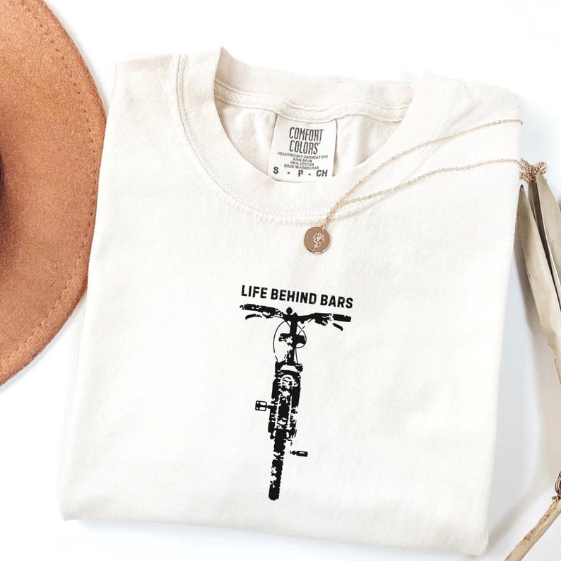 May include: Cream-colored t-shirt with the words "LIFE BEHIND BARS" above a black bicycle illustration. The shirt has a crew neck and short sleeves. A gold necklace with a pendant is draped across the shirt.