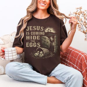 May include: A brown t-shirt with the text "JESUS IS COMING HIDE THE EGGS" in a vintage style font. The graphic includes an image of Jesus, a tomb, a basket of eggs, and three crosses. The shirt is a crew neck.