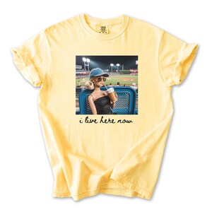 Pu&ograve; includere: T-shirt giallo chiaro con una grafica di una bambola con occhiali da sole e berretto da baseball, che tiene in mano una bevanda a una partita di baseball. Il testo "i live here now" &egrave; sotto.