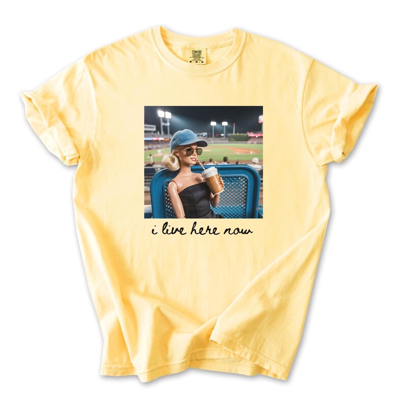 Pu&ograve; includere: T-shirt giallo chiaro con una grafica di una bambola con occhiali da sole e berretto da baseball, che tiene in mano una bevanda a una partita di baseball. Il testo "i live here now" &egrave; sotto.