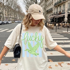 May include: Cream-colored t-shirt with the word "lucky" in green script and a green duck graphic. The shirt has blue vertical stripes behind the duck. A beige baseball cap and a black purse with a chain strap are also visible.
