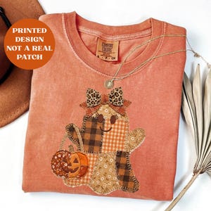 May include: A terracotta-colored crewneck t-shirt featuring a printed design of a ghost with a leopard print bow, holding a pumpkin. The ghost is composed of various brown, orange, and cream plaid and floral patterns. Text reads: PRINTED DESIGN NOT A REAL PATCH.