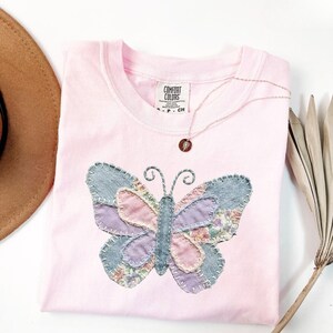 May include: A pale pink Comfort Colors t-shirt with a butterfly applique. The butterfly is made of denim and floral fabric in shades of blue, pink, and purple. A gold necklace with a pendant is also visible.