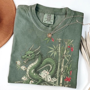May include: Green Comfort Colors t-shirt featuring a detailed illustration of a green dragon with mahjong tiles, bamboo, and red flowers. A silver necklace with a pendant rests on the shirt. The shirt is folded.