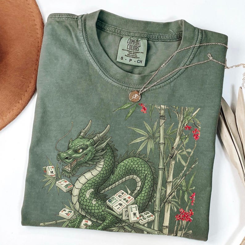 May include: Green Comfort Colors t-shirt featuring a detailed illustration of a green dragon with mahjong tiles, bamboo, and red flowers. A silver necklace with a pendant rests on the shirt. The shirt is folded.