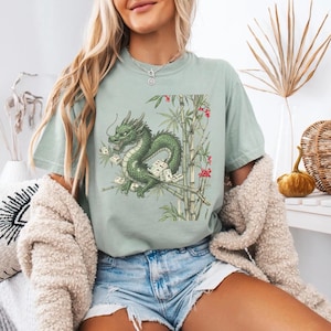 May include: A light green t-shirt featuring a detailed illustration of a green dragon perched on bamboo stalks. The dragon is surrounded by Mahjong tiles and red flowers. The shirt has a relaxed fit.