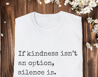 May include: White t-shirt with the text "If kindness isn't an option, silence is" in black. The shirt is folded on a wooden surface, with white flowers in the background.