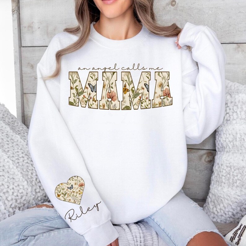 Pode incluir: Camisola branca com o texto "an angel calls me MAMA" em um design floral. A manga apresenta um design floral em forma de cora&ccedil;&atilde;o com o nome "Riley" em escrita cursiva. Um top aconchegante e elegante.