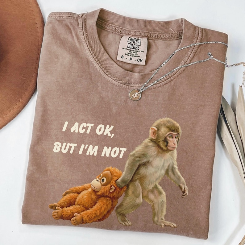 May include: A brown Comfort Colors t-shirt with the text "I ACT OK, BUT I'M NOT" and a graphic of two monkeys. One monkey is walking, pulling the other. A silver necklace with a pendant is on the shirt.