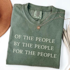 May include: An olive green t-shirt with the quote "OF THE PEOPLE BY THE PEOPLE FOR THE PEOPLE" printed in white. A gold-colored necklace with a circular pendant rests on the shirt. The shirt is folded neatly.