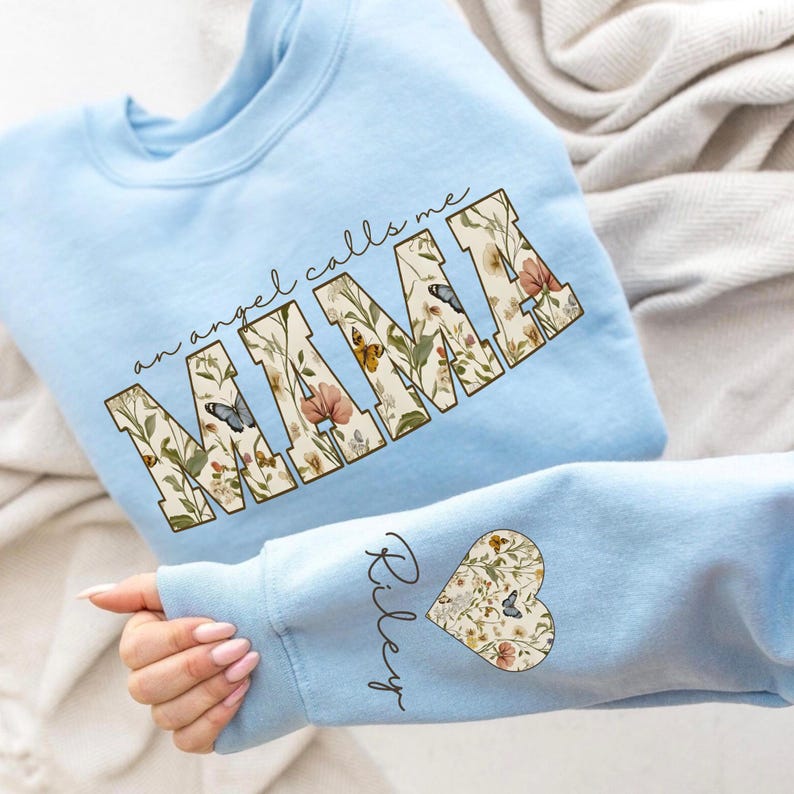 Pode incluir: Camisola azul clara com o texto "an angel calls me MAMA" num design floral. A manga apresenta um design floral em forma de cora&ccedil;&atilde;o e o nome "Riley". A camisola &eacute; feita de um material macio.