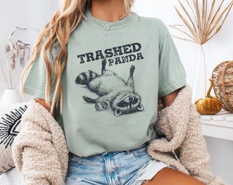 Könnte beinhalten: Hellgrünes T-Shirt mit dem Text "Trashed Panda" und einer Grafik eines auf dem Rücken liegenden Waschbären, der eine Bierdose hält. Das T-Shirt hat eine lockere Passform.