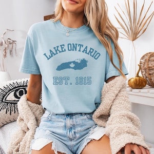 May include: A light blue short-sleeve t-shirt featuring dark blue text that reads LAKE ONTARIO and EST. 1615, with a central graphic of the lake silhouette containing a small white maple leaf icon. The shirt has a crew neckline and a relaxed fit.