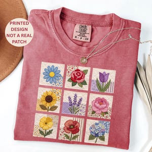 Peut inclure: Un t-shirt Comfort Colors rose poussiéreux avec un motif floral imprimé. Le motif comprend neuf carrés, chacun présentant une fleur différente, comme un tournesol, une rose et une tulipe. Un collier en argent avec un pendentif repose sur le t-shirt.