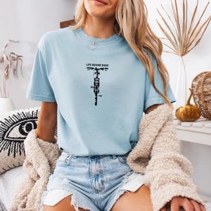 May include: Light blue t-shirt with the text "LIFE BEHIND BARS" above a black bicycle graphic. The shirt is a short-sleeved crew neck style. The model is wearing denim shorts and a cream-colored sweater.