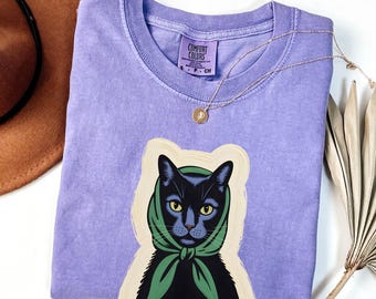 May include: A lilac Comfort Colors t-shirt featuring a graphic of a black cat wearing a green scarf. The cat has yellow eyes and is set against a cream-colored background. A gold necklace is draped across the shirt.