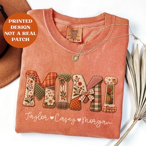 May include: A terracotta-coloured t-shirt featuring a printed design of the word MIMI in a patchwork style with floral, plaid, and polka dot patterns. Below the word, the names Taylor, Casey, and Morgan are printed in white script with hearts.