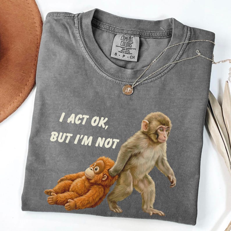 May include: A gray Comfort Colors t-shirt with the text "I ACT OK, BUT I'M NOT" and a graphic of two monkeys. One monkey is pulling the other. A silver necklace with a pendant is on the shirt.