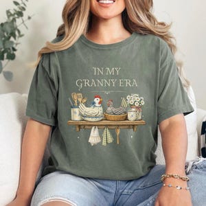 May include: Sage green t-shirt with the text "IN MY GRANNY ERA". The graphic features a shelf with chickens, kitchen utensils, and flowers. Two decorative towels and a wooden spoon hang below the shelf. The shirt is a comfortable, casual style.