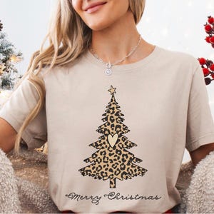 May include: Beige t-shirt with a Christmas tree design in a leopard print pattern, a heart, and a star. The words "Merry Christmas" are written in a script font. A festive holiday tee, perfect for the Christmas season.