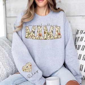 Pode incluir: Um moletom cinza claro com o texto "an angel calls me MAMA" em um design floral. Um design floral em forma de cora&ccedil;&atilde;o com o nome "Riley" est&aacute; na manga. O moletom &eacute; um estilo casual.