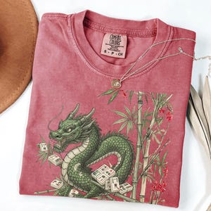 May include: A red-pink Comfort Colors t-shirt featuring a green dragon design with bamboo and Mahjong tiles. The shirt has a crew neck and short sleeves. A silver necklace with a pendant is draped across the shirt.