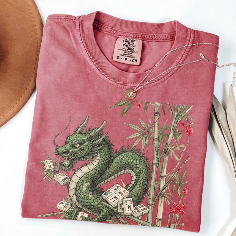 May include: A red-pink Comfort Colors t-shirt featuring a green dragon design with bamboo and Mahjong tiles. The shirt has a crew neck and short sleeves. A silver necklace with a pendant is draped across the shirt.