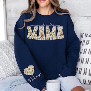 Pode incluir: Camisola azul marinho com o texto "an angel calls me MAMA". A palavra "MAMA" &eacute; preenchida com um desenho floral e borboletas. Um desenho floral em forma de cora&ccedil;&atilde;o com o nome "Riley" est&aacute; na manga.