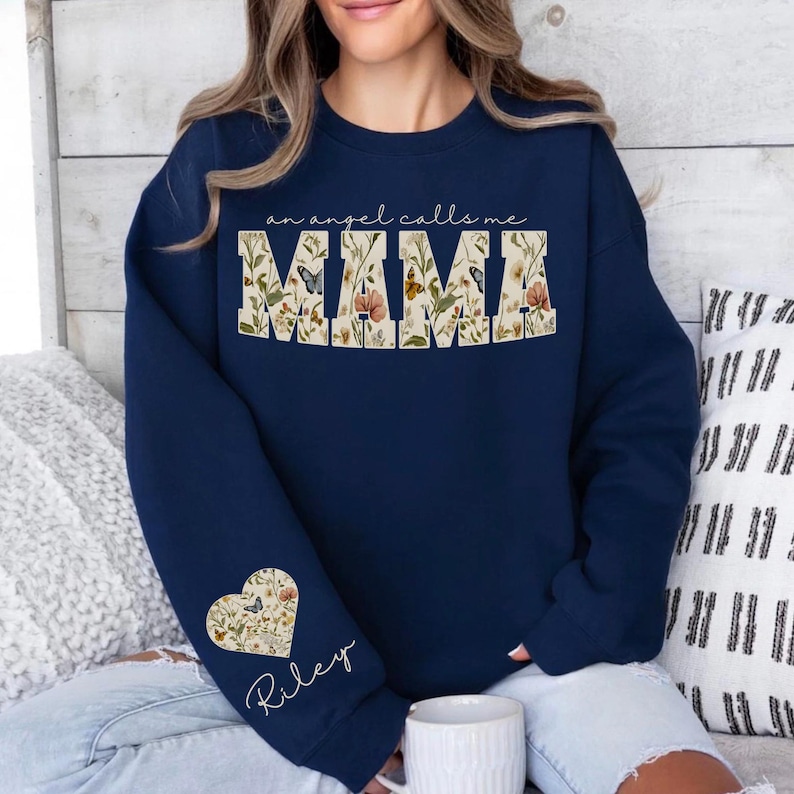 Pode incluir: Camisola azul marinho com o texto "an angel calls me MAMA". A palavra "MAMA" &eacute; preenchida com um desenho floral e borboletas. Um desenho floral em forma de cora&ccedil;&atilde;o com o nome "Riley" est&aacute; na manga.