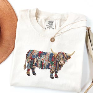 Könnte beinhalten: Cremefarbenes T-Shirt mit einem farbenfrohen Patchwork-Design einer Highland-Kuh. Die Kuh hat lange Hörner und besteht aus verschiedenen Stoffmustern in Rot, Blau und Grün. Das Shirt hat ein Comfort Colors-Etikett.