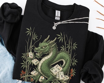 May include: A black long-sleeve sweatshirt featuring a graphic print of a green dragon surrounded by bamboo stalks and scattered mahjong tiles. The sweatshirt has a crew neckline and a Gildan brand label visible at the inner collar area.