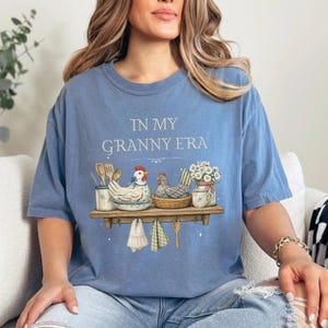 May include: A light blue t-shirt with the text "IN MY GRANNY ERA". The shirt features a graphic of a shelf with chickens, flowers, and kitchen utensils. The design includes a white and blue color scheme.