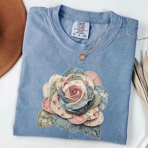 Peut inclure: Un t-shirt bleu Comfort Colors avec une grande application de rose en tissu. La rose présente des couches de pétales roses, bleus et à motifs. Un collier en or avec un pendentif repose sur le t-shirt. Taille S-P-CH.