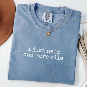 May include: A blue Comfort Colors t-shirt with the text "i just need one more tile" in white. A gold necklace with a pendant rests on the shirt. The shirt is folded and has a small tag.