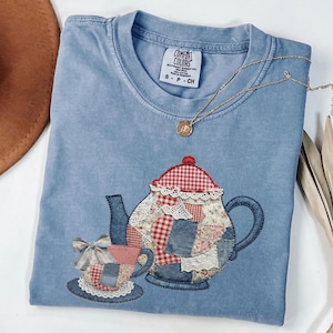 May include: A blue Comfort Colors t-shirt featuring an embroidered patchwork design of a teapot and teacup. The teapot has a red and white checkered lid and floral patches. A gold necklace with a pendant rests on the shirt.