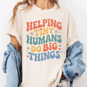 Può includere: T-shirt color crema con la scritta "Helping Tiny Humans Do Big Things" in un font retrò. Il testo è nei toni dell'arancione, giallo, verde acqua e blu, con decorazioni floreali e a forma di cuore. La maglietta ha maniche corte.