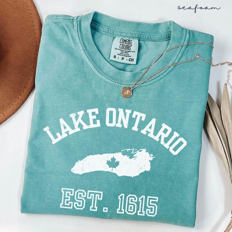May include: A seafoam green short-sleeve t-shirt with a crew neckline. The shirt features white screen-printed text that reads LAKE ONTARIO, a graphic outline of the lake with a maple leaf, and the text EST. 1615. A gold-toned necklace rests on the shirt.