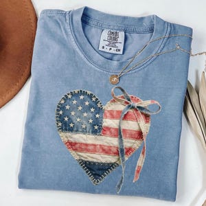 May include: A blue Comfort Colors t-shirt featuring a heart-shaped American flag design. The flag is made of denim and red and white striped fabric, with a blue bow. A gold necklace with a pendant rests on the shirt.
