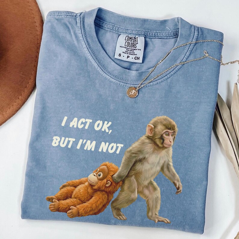 May include: A blue Comfort Colors t-shirt with the text "I ACT OK, BUT I'M NOT" and an illustration of two monkeys. One monkey is pulling the other. A gold necklace is draped across the shirt.