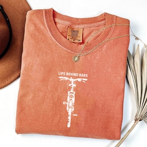 May include: A coral-colored t-shirt with a white motorcycle graphic and the text "LIFE BEHIND BARS." A gold necklace with a pendant rests on the shirt. The shirt is folded, with a brown hat visible in the background.