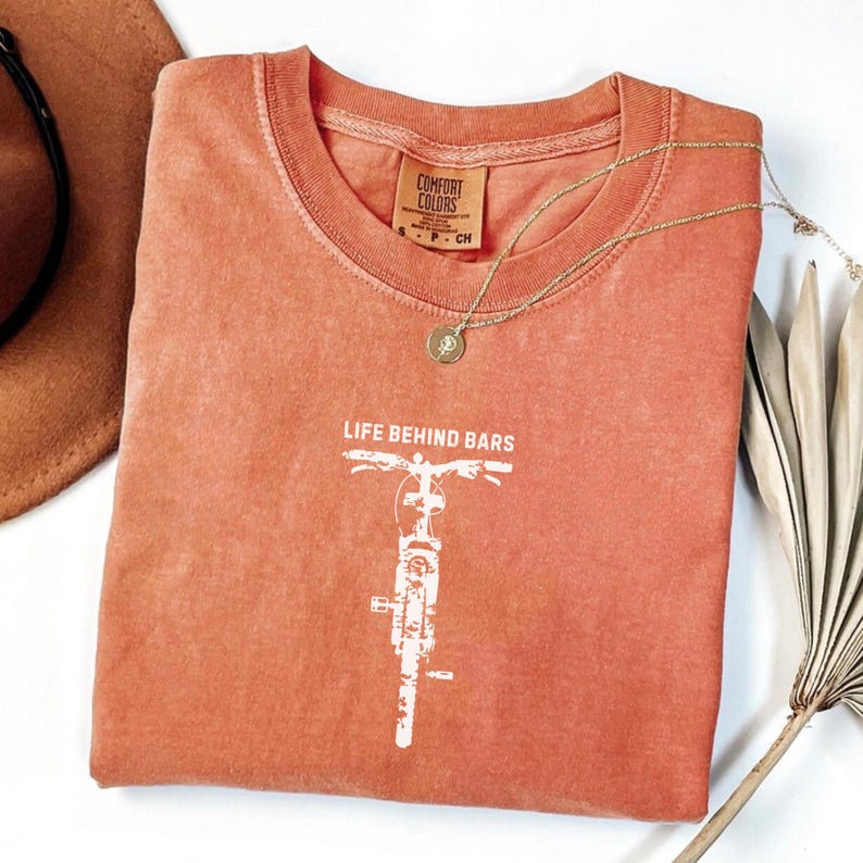 May include: A coral-colored t-shirt with a white motorcycle graphic and the text "LIFE BEHIND BARS." A gold necklace with a pendant rests on the shirt. The shirt is folded, with a brown hat visible in the background.