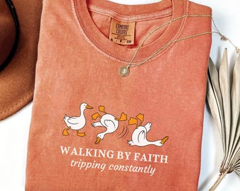 May include: A coral-colored Comfort Colors t-shirt featuring a graphic of three white ducks with orange feet, one tripping. The text "WALKING BY FAITH tripping constantly" is printed below the ducks. A gold necklace with a pendant is draped on the shirt.