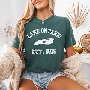 May include: A dark green short-sleeve t-shirt featuring white text that reads LAKE ONTARIO and EST. 1615. Between the text is a white graphic outline of Lake Ontario with a small maple leaf icon centered within the shape of the lake.